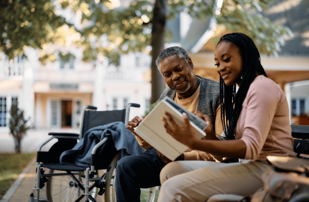 A senior and adult child sit on a park bench looking through a photo album together.