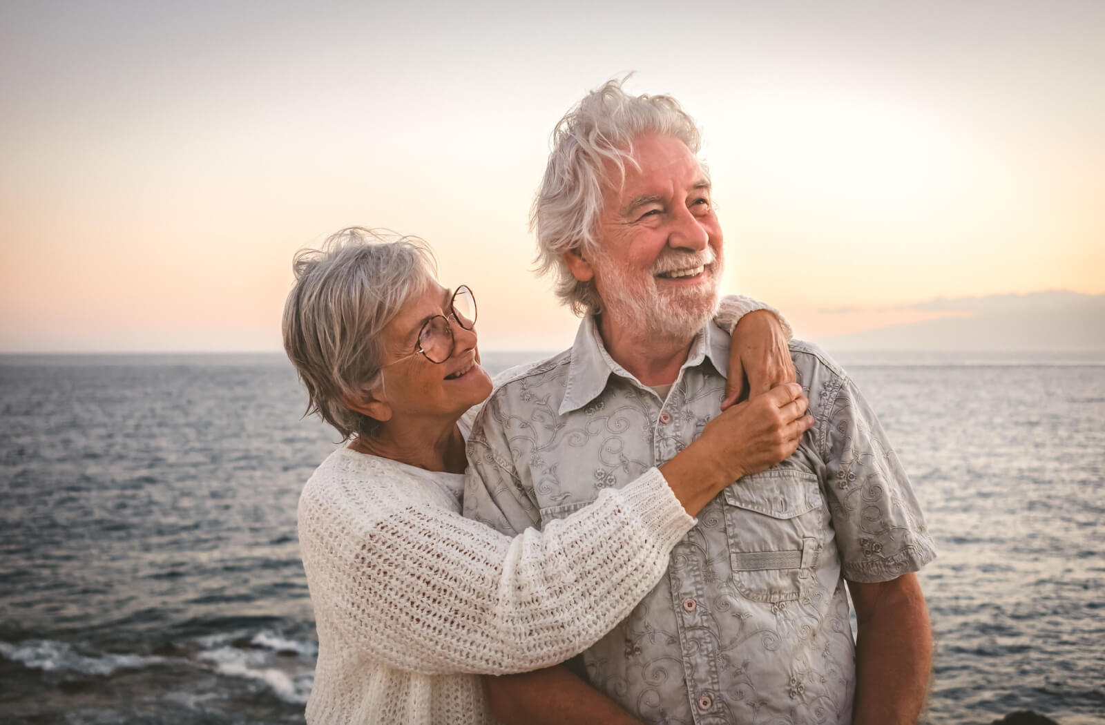 A senior woman and her husband smiling and hugging on a day trip to the beach at sunset.