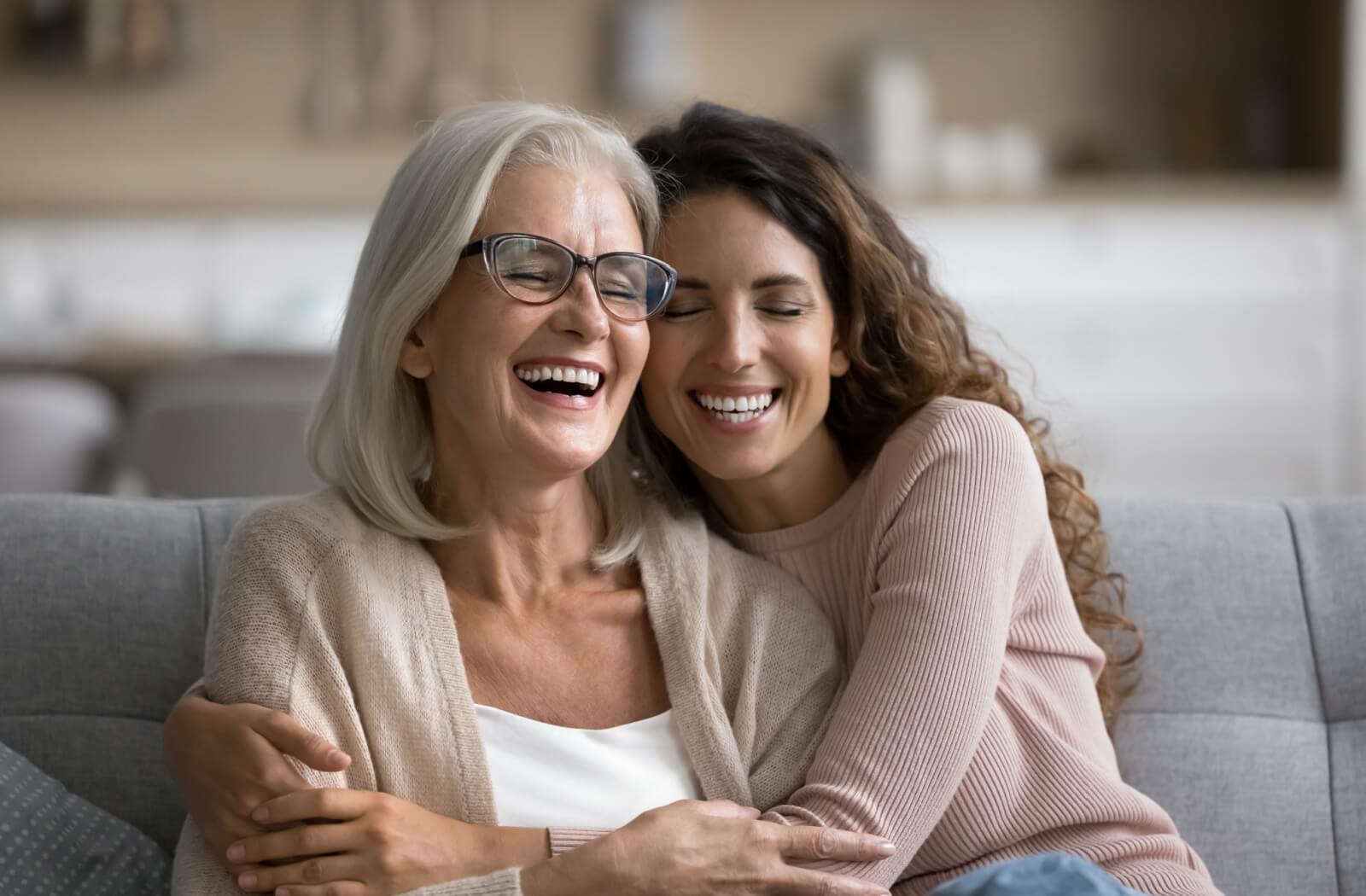 A happy senior and their adult child enjoying a warm embrace as they sit on a couch and chat.