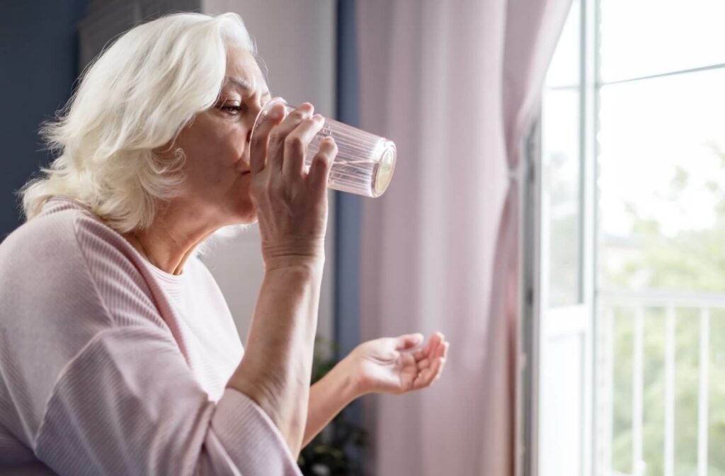 Older adult drinking a glass of water while standing by a bright window with curtains in the background