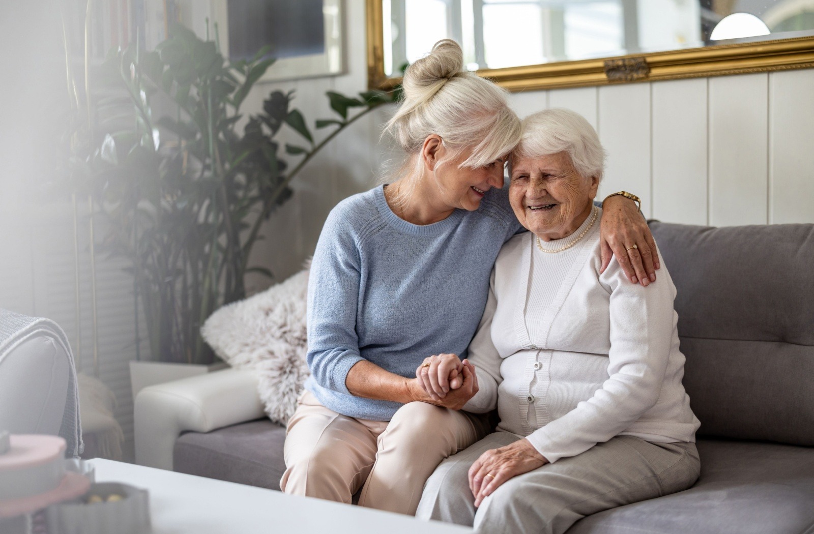 A smiling caregiver hugs their senior loved one on a couch in a bright living room.