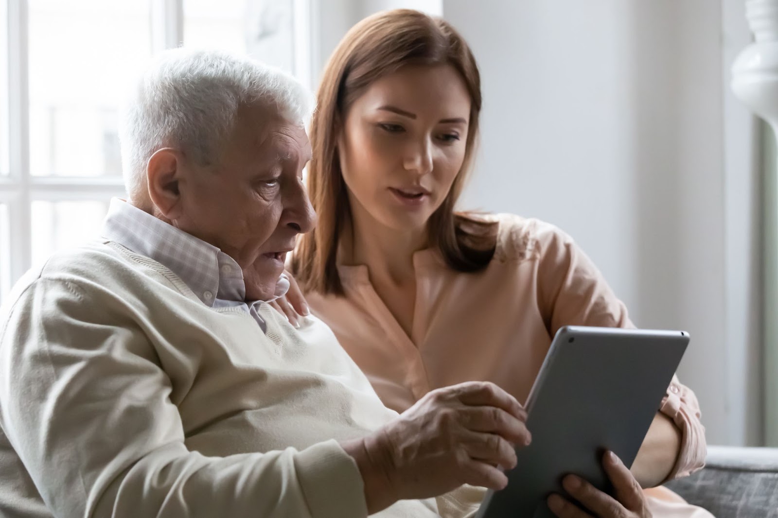 a younger person sits on a couch with a senior, they are both looking at a tablet